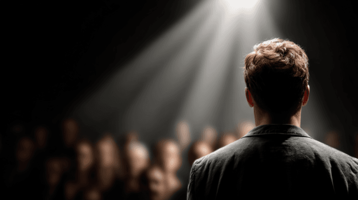 Person speaking on stage, spotlighted audience listening.