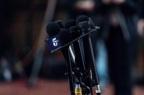 A cluster of news microphones mounted on a stand at a press conference, one carrying a TV station flag