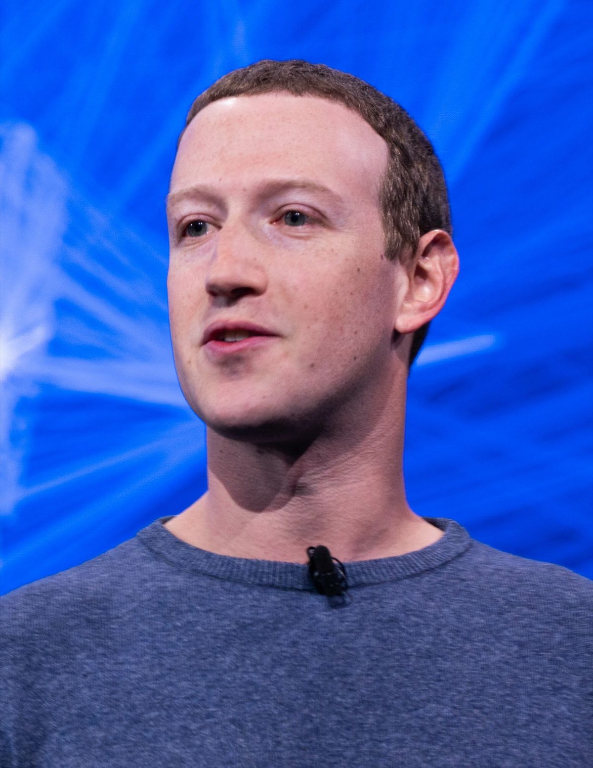 Mark Zuckerberg in a gray sweater speaking on stage in front of a bright blue backdrop