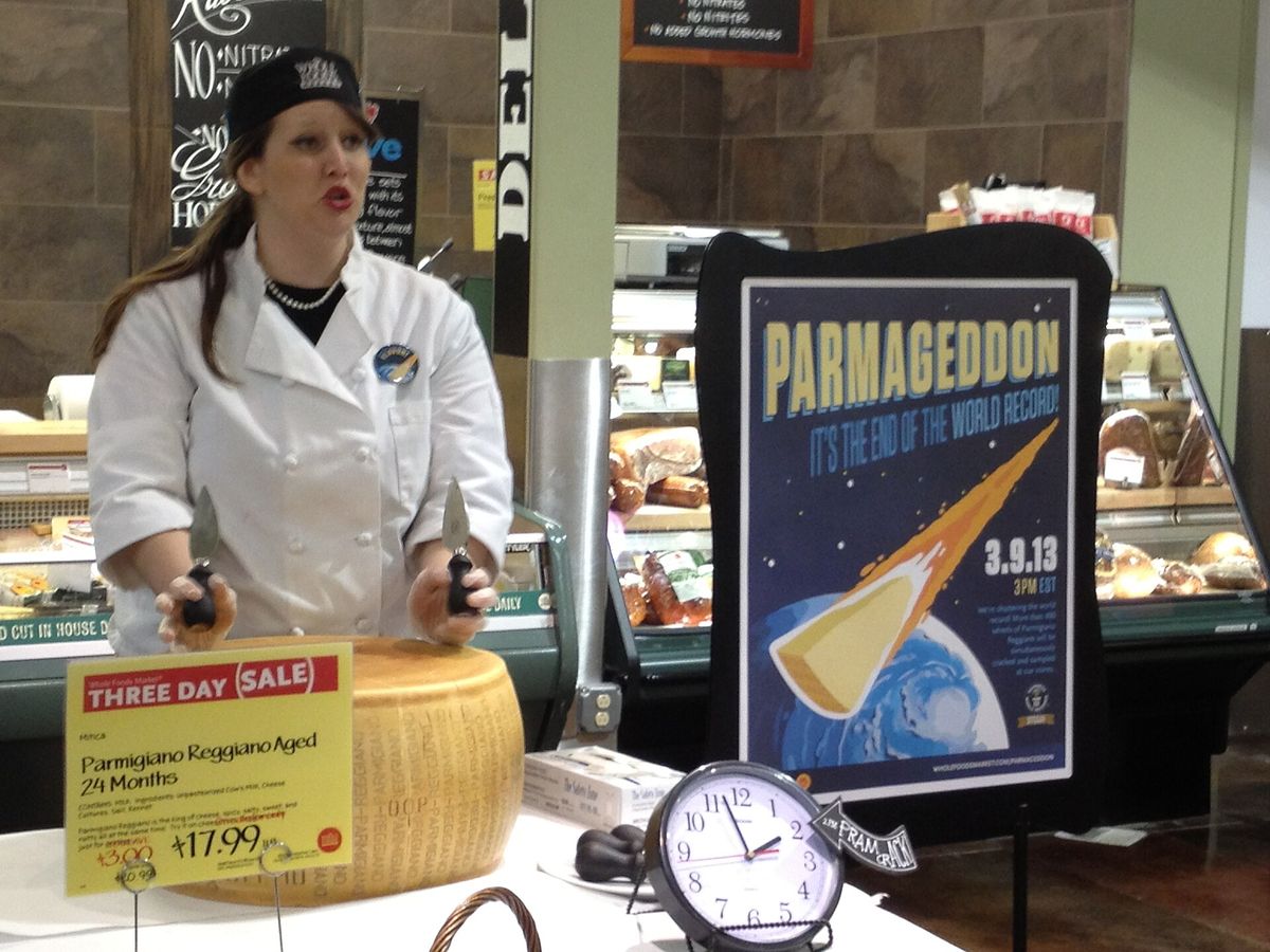 A woman in a white chef's jacket holds two cheese knives over a large wheel of Parmigiano Reggiano, beside a sale sign and a poster reading Parmageddon