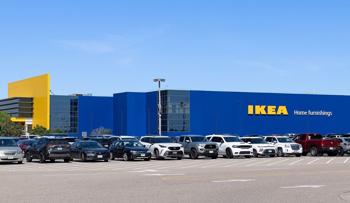 A long blue IKEA store with a yellow entrance block and a large parking lot full of cars and pickup trucks under a clear blue sky