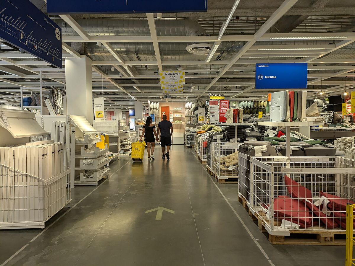 A wide aisle inside an IKEA store with a yellow arrow on the floor, white shelving on the left, wire bins of cushions and textiles on the right, and two shoppers walking away pulling a yellow bag