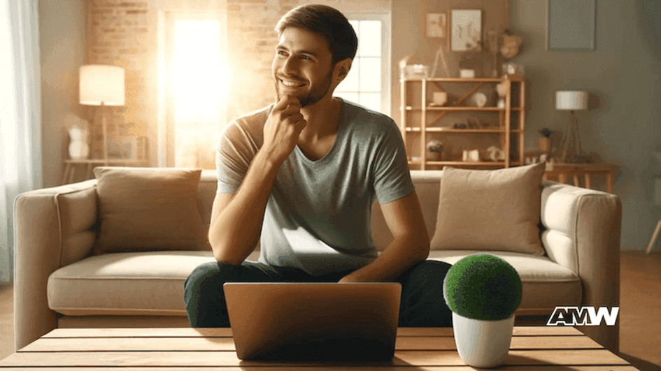 A smiling man sitting on a sofa behind an open laptop in a sunlit living room, resting his chin on his hand