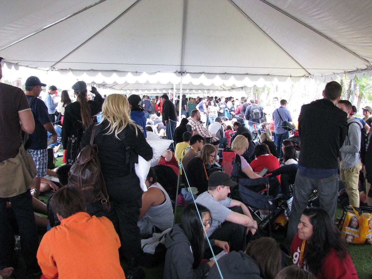 A crowd of fans sitting and standing on grass under a large white tent, waiting in line at San Diego Comic-Con