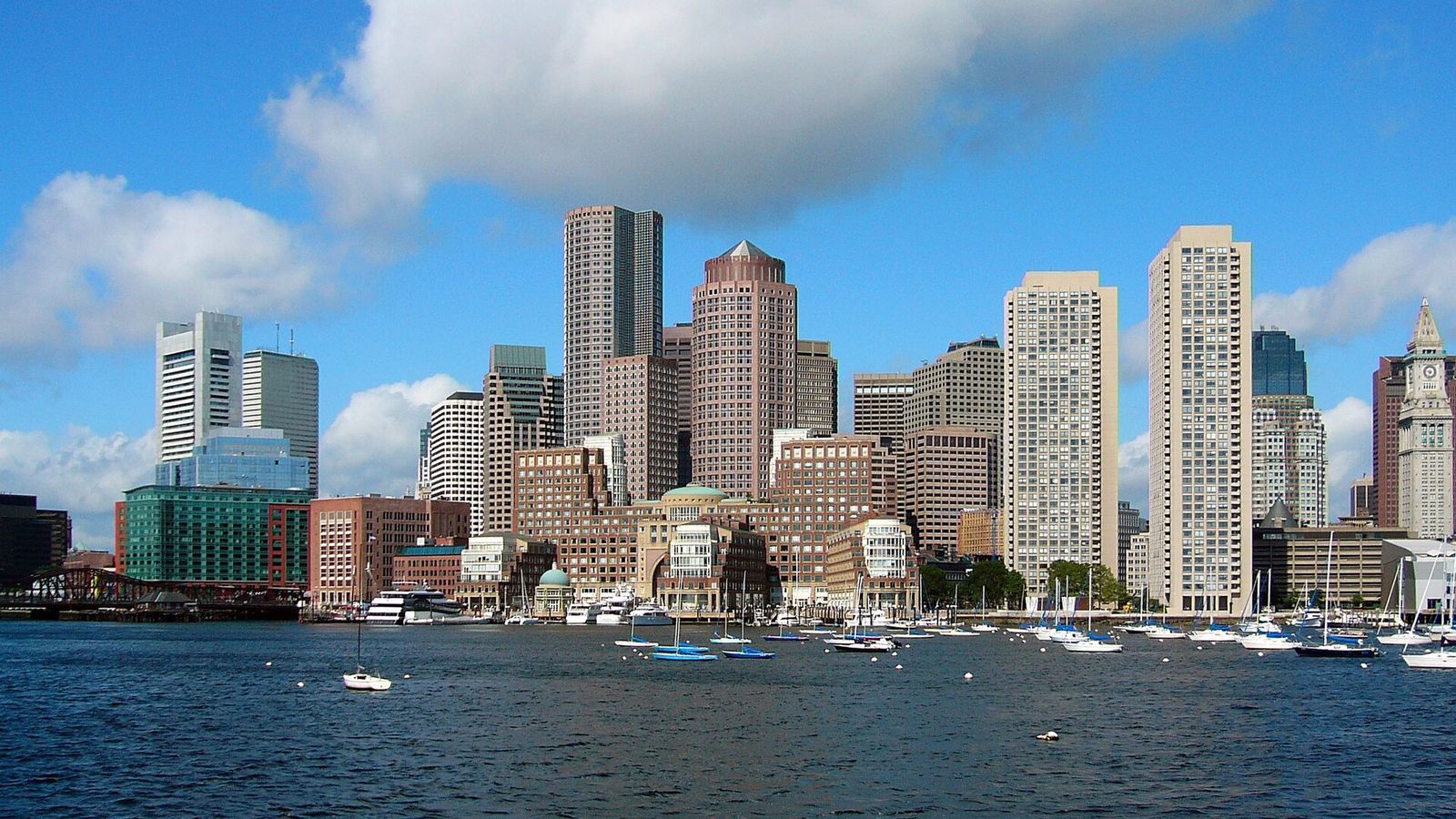 The Boston Financial District skyline seen from the harbor, with high-rise office towers behind a marina of moored sailboats under a large white cloud