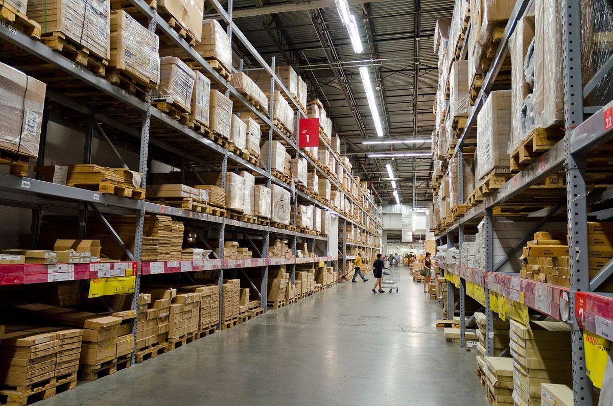 A long warehouse aisle in an IKEA store lined with tall metal shelving holding flat-packed boxes, with a few shoppers far down the aisle