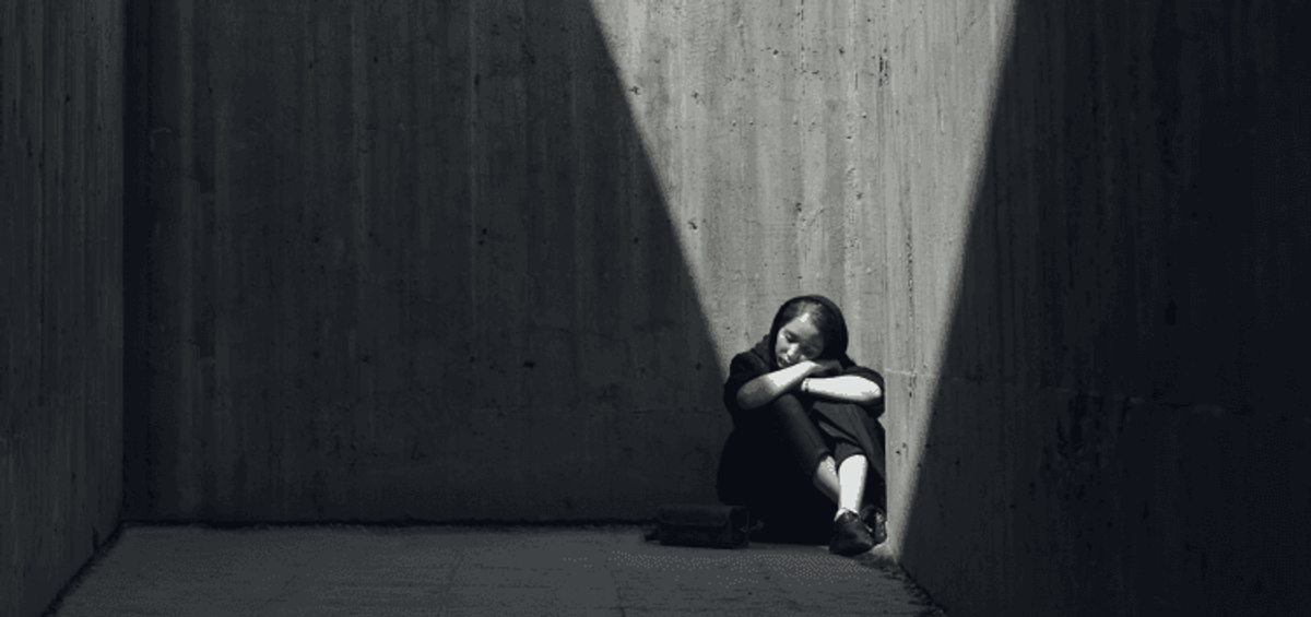 Person sitting alone in dark concrete corner.