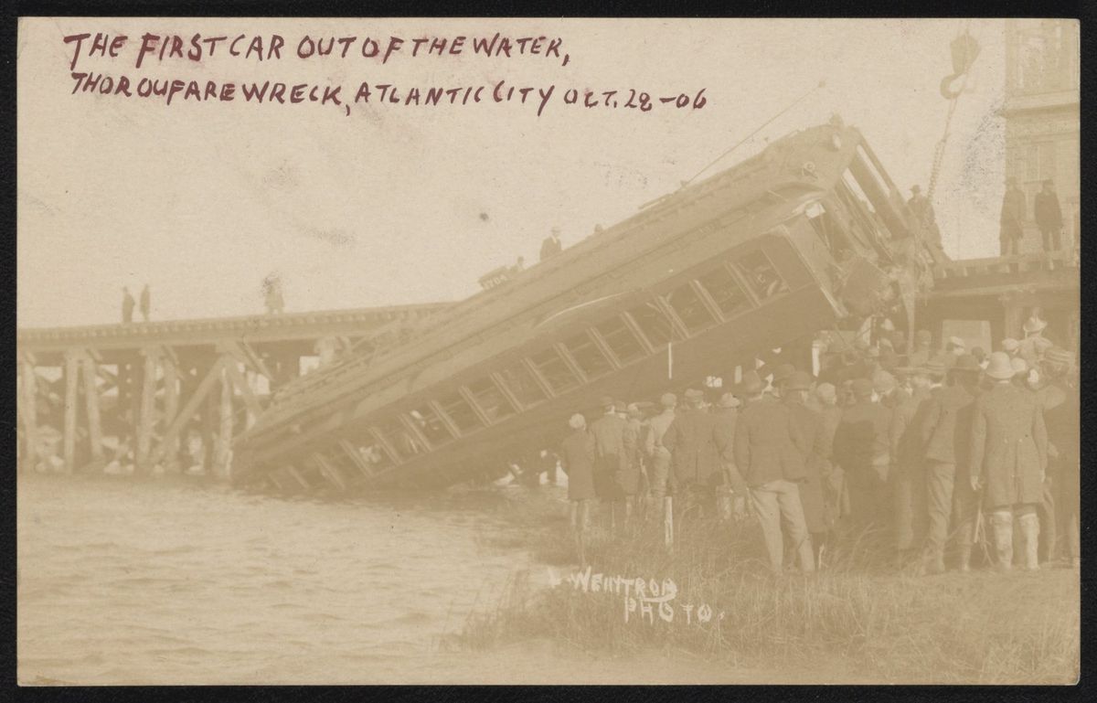 Sepia postcard photograph of a railroad coach being hoisted at a steep angle out of the water beside a trestle while a large crowd in hats and coats watches from the bank, captioned "The first car out of the water, Thorofare wreck, Atlantic City"