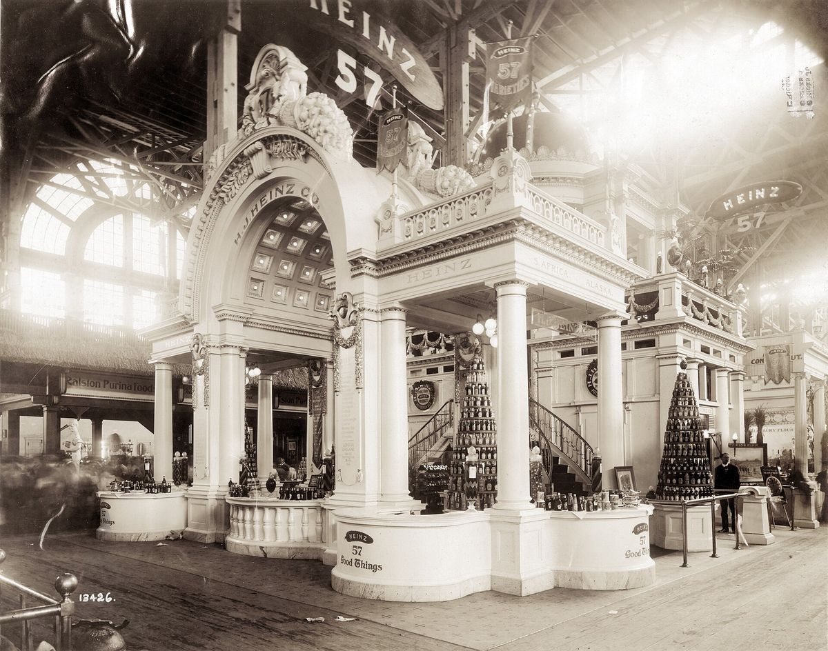 Black and white photograph of an ornate white Heinz exhibit booth with columns, an arched entrance, pyramids of bottled products and signs reading Heinz 57 and 57 Good Things inside a large exhibition hall