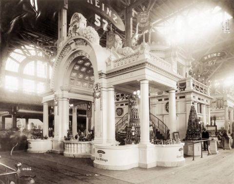 Black and white photograph of an ornate white Heinz exhibit booth with columns, an arched entrance, pyramids of bottled products and signs reading Heinz 57 and 57 Good Things inside a large exhibition hall