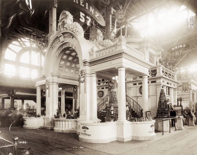Black and white photograph of an ornate white Heinz exhibit booth with columns, an arched entrance, pyramids of bottled products and signs reading Heinz 57 and 57 Good Things inside a large exhibition hall