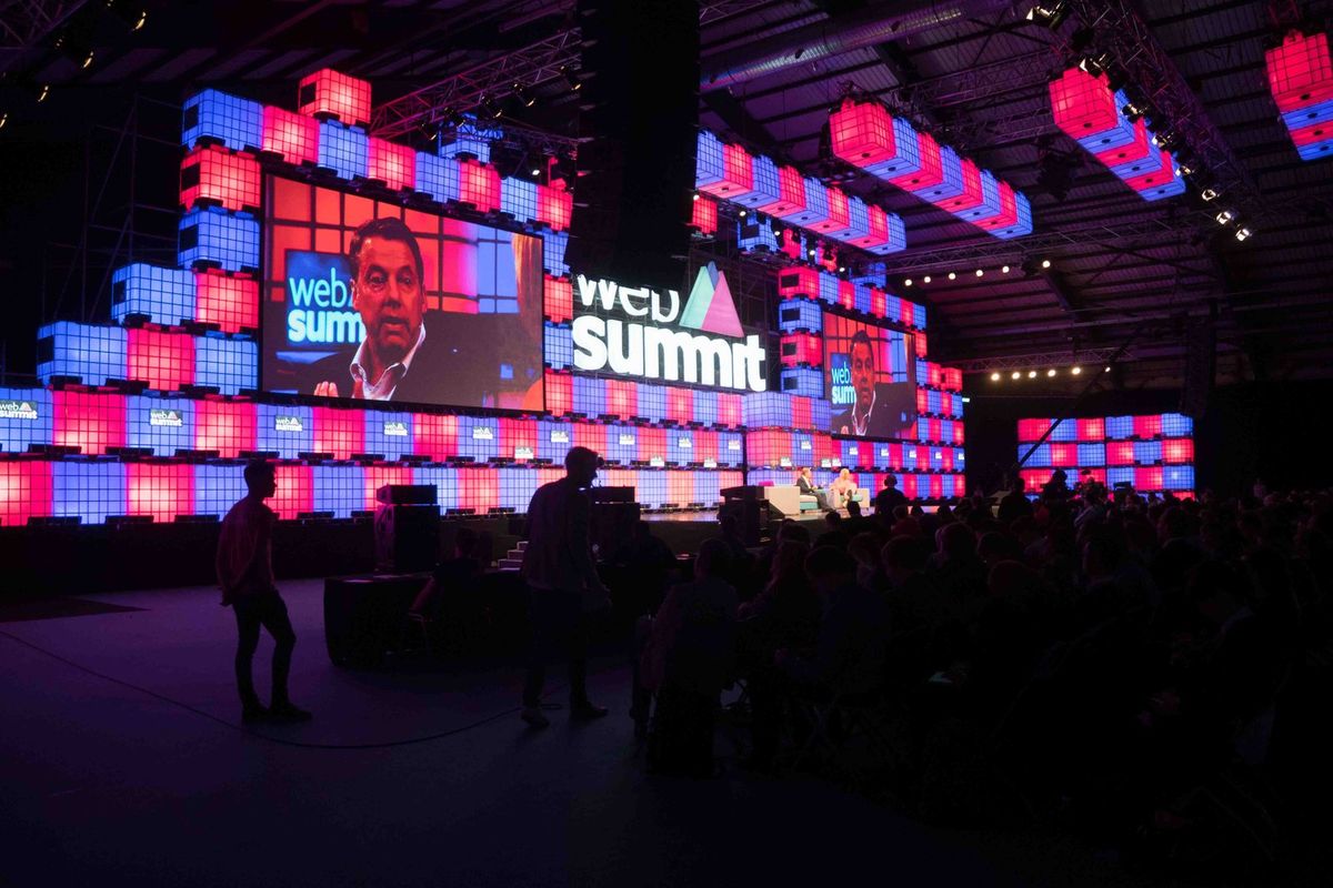 A dark arena with a large audience in silhouette facing a stage built from glowing red and blue cubes, with a speaker shown on big screens beside the Web Summit logo