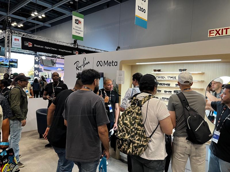 Visitors crowding a Ray-Ban and Meta booth at a trade show, with staff in Meta shirts and a wall of smart glasses on display shelves