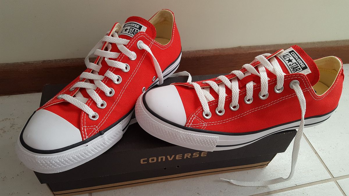 A pair of red low-top Converse All Star sneakers with white toe caps and laces, standing on a black Converse shoe box on a tiled floor