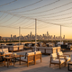 Premium outdoor event terrace at golden hour with elegant furniture, string lights, and city skyline