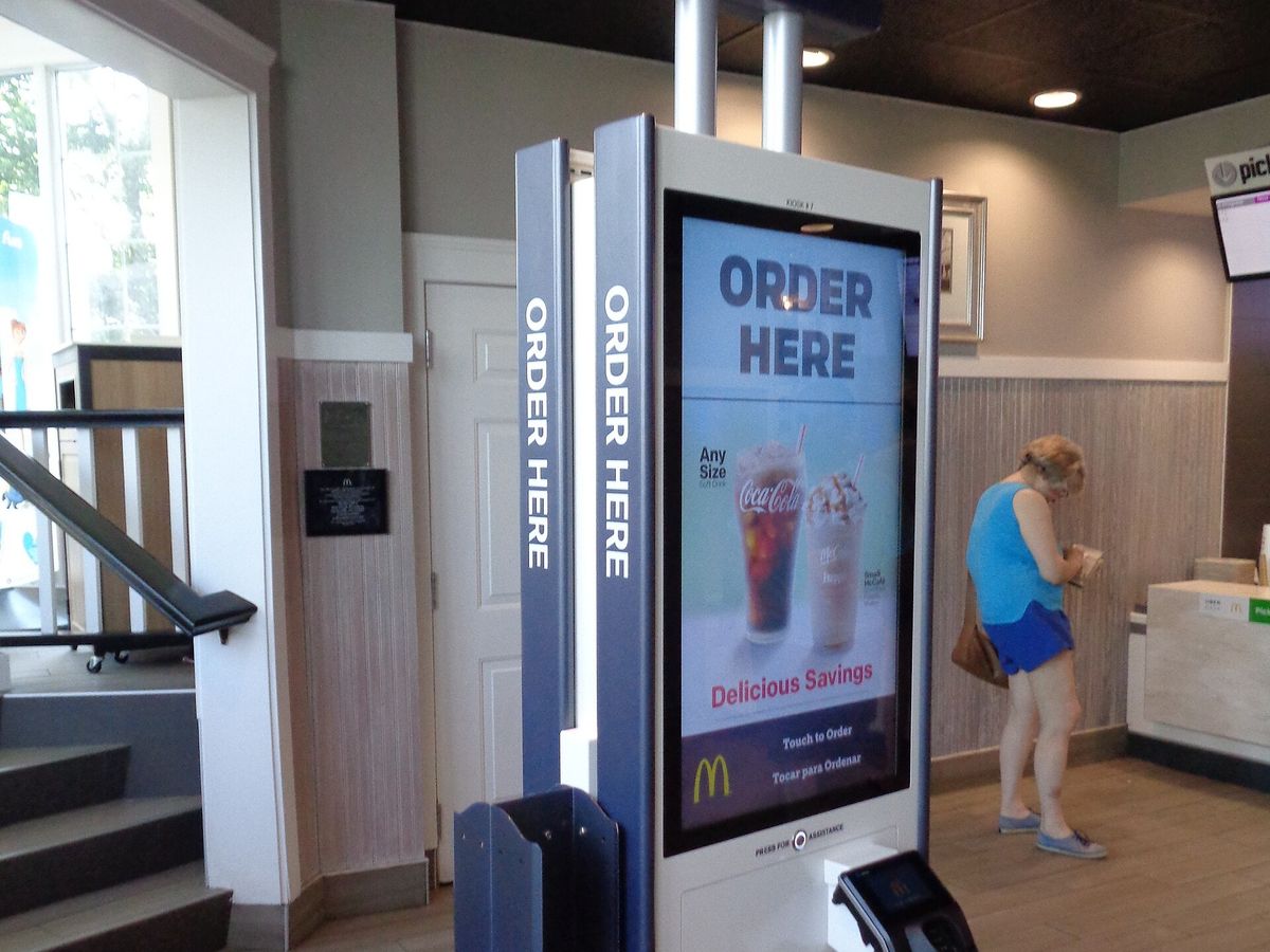 A self-order kiosk labeled Order Here inside a McDonald's restaurant, its screen showing an iced soft drink and a frappé, with a customer standing at the front counter in the background