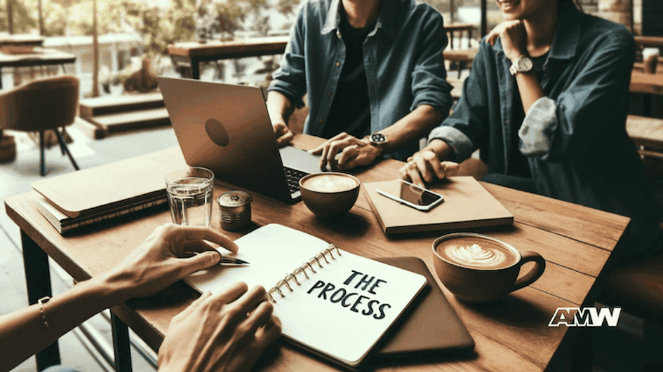 Two people sitting at a wooden café table with a laptop, a phone and an open spiral notebook with the words The Process written on the page