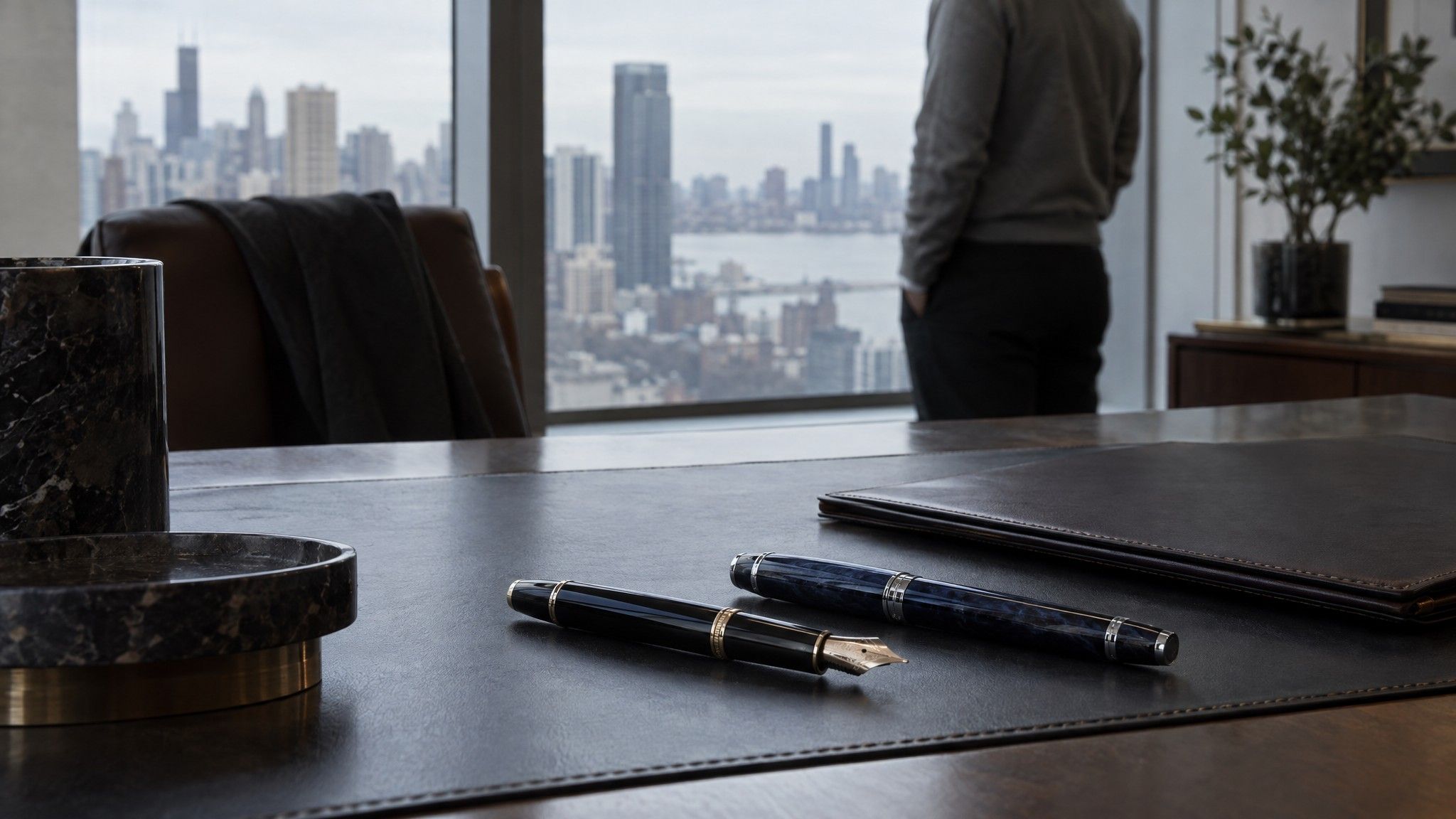 Two fountain pens on a leather desk blotter beside a closed folder
