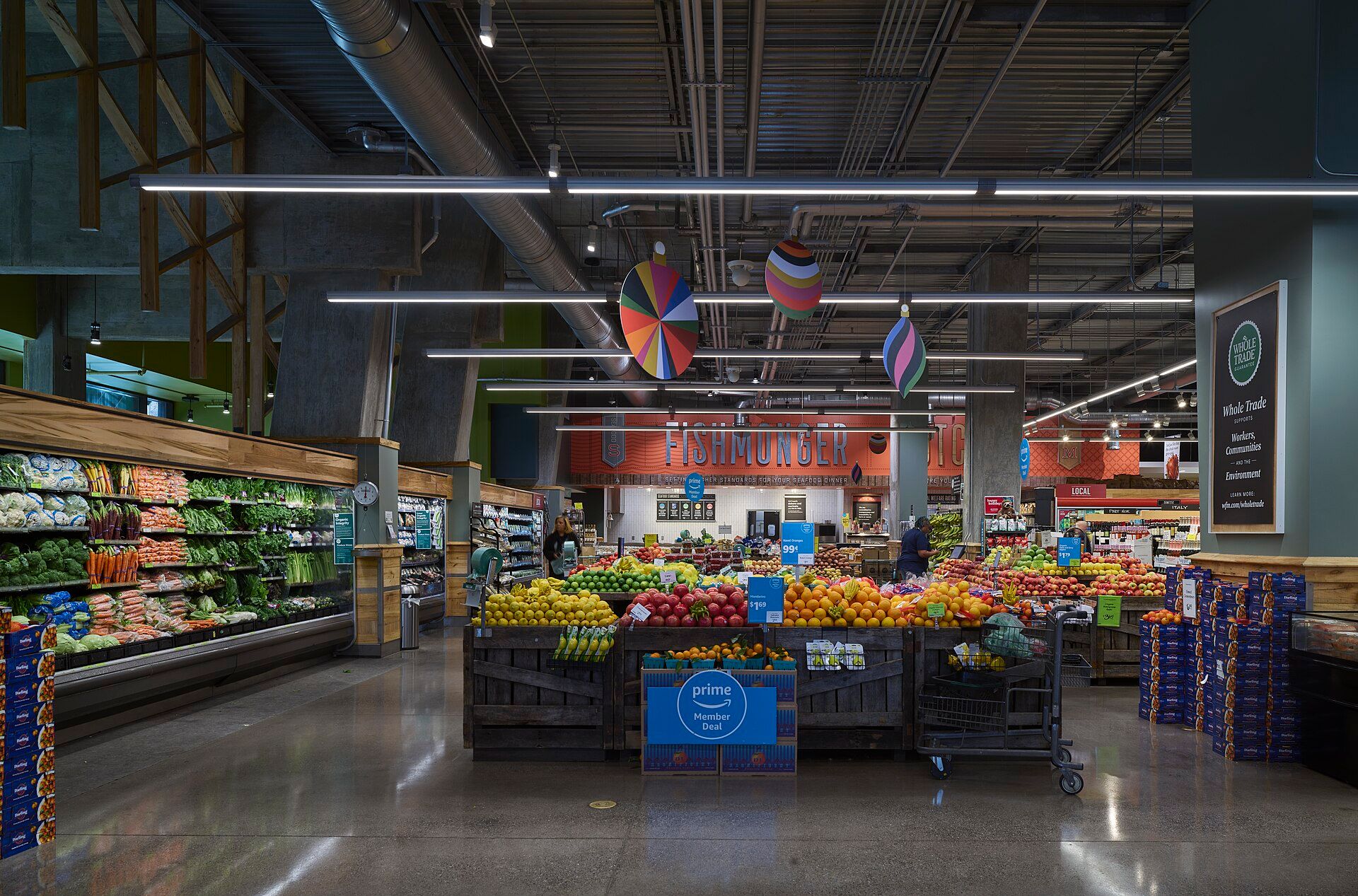 Produce section of a Whole Foods Market with fruit stacked on wooden crates, a blue Prime Member Deal sign, a Whole Trade sign and a Fishmonger counter at the back