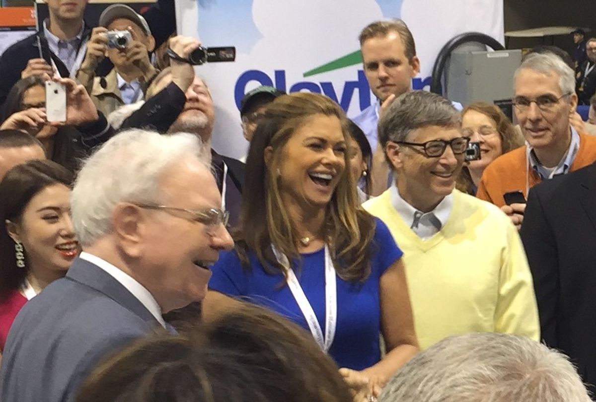 Warren Buffett smiling in the foreground as Kathy Ireland and Bill Gates laugh beside him, surrounded by shareholders holding up phones and cameras in the exhibition hall