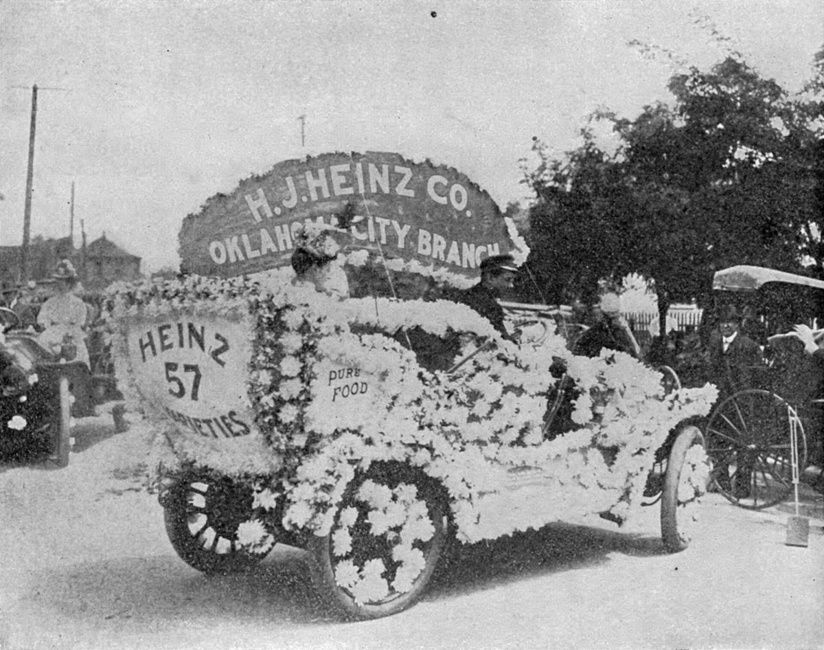 Black and white photograph of an early automobile covered in white flowers with a sign reading H. J. Heinz Co. Oklahoma City Branch and panels reading Heinz 57 Varieties and Pure Food, with people seated inside, on a street lined with trees and other carriages