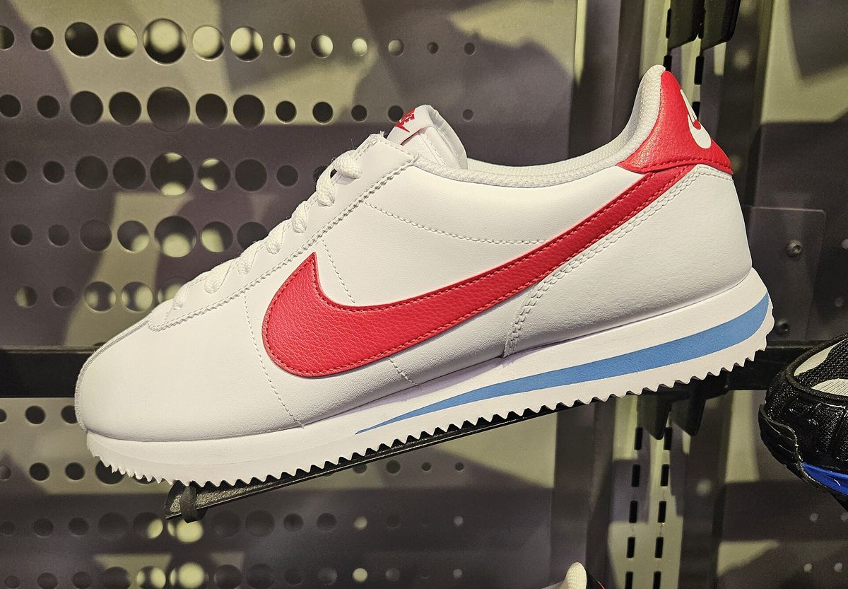 A white leather Nike Cortez with a red swoosh and a blue stripe along the sole, displayed on a perforated metal shelf