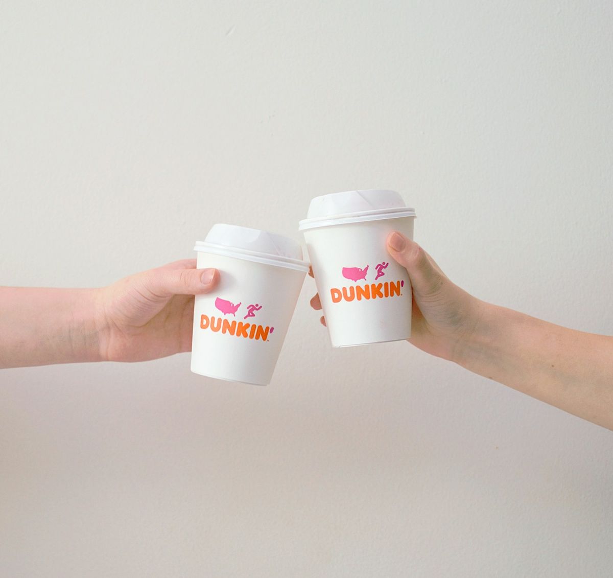 Two hands raising white Dunkin' paper cups with lids toward each other against a plain white wall
