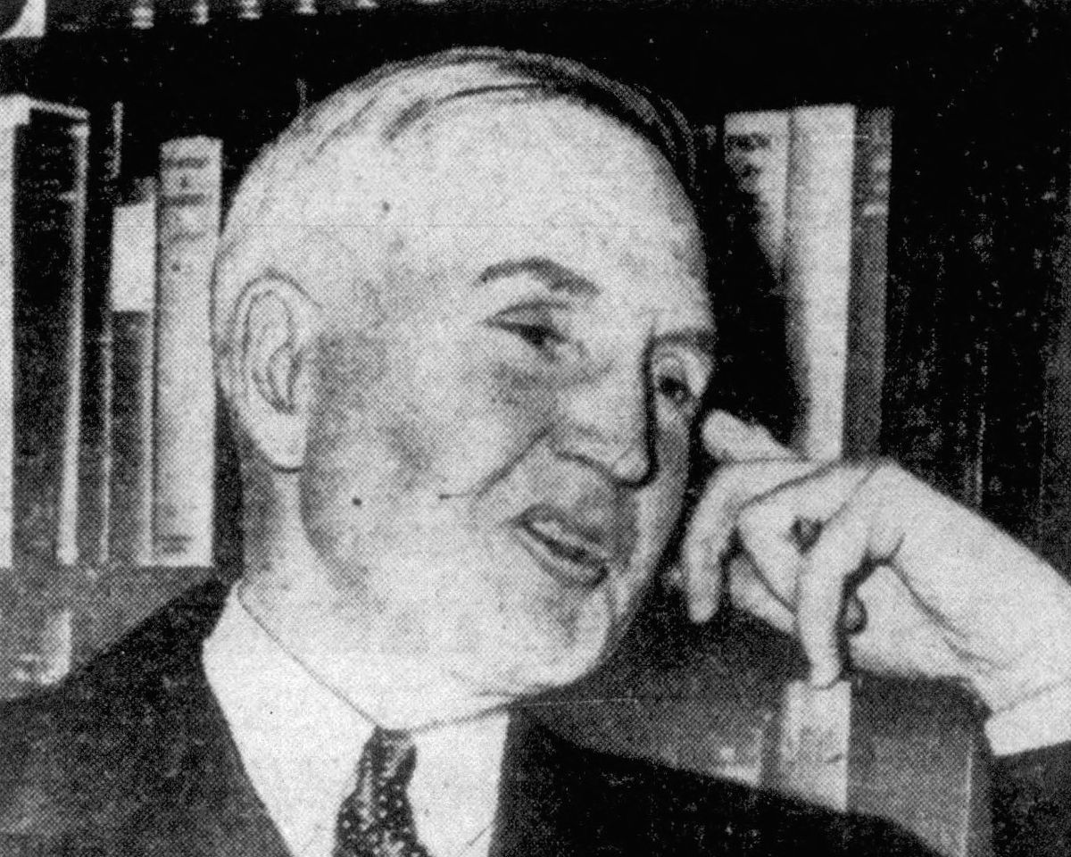 Black-and-white newspaper photograph of Sewell Avery, an elderly man with a short white beard, suit and polka-dot tie, fingers raised to his face in front of a bookshelf