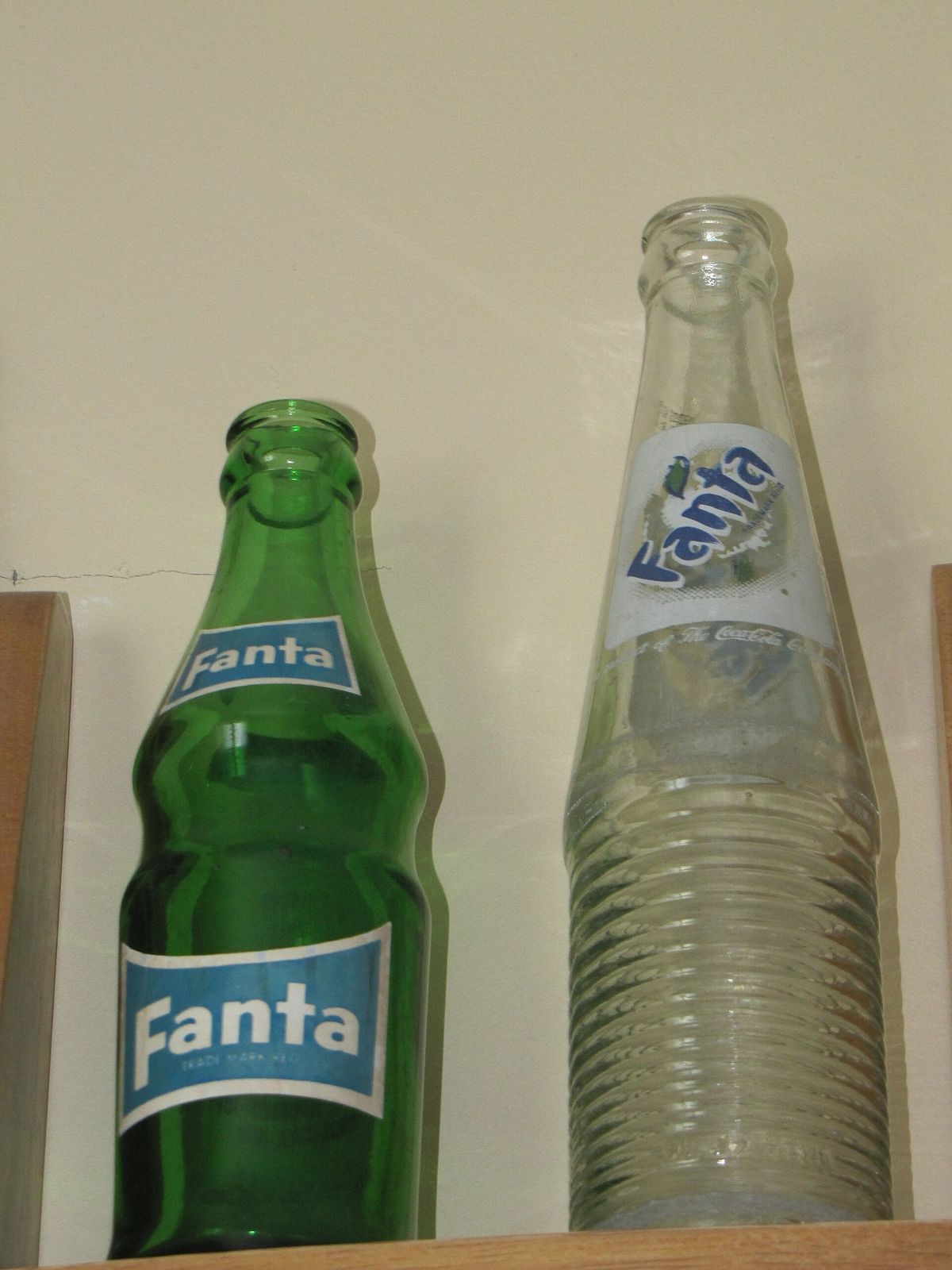 Two old Fanta glass bottles on a wooden shelf, a green bottle with a blue Fanta label and a clear bottle with horizontal ridges and a printed logo