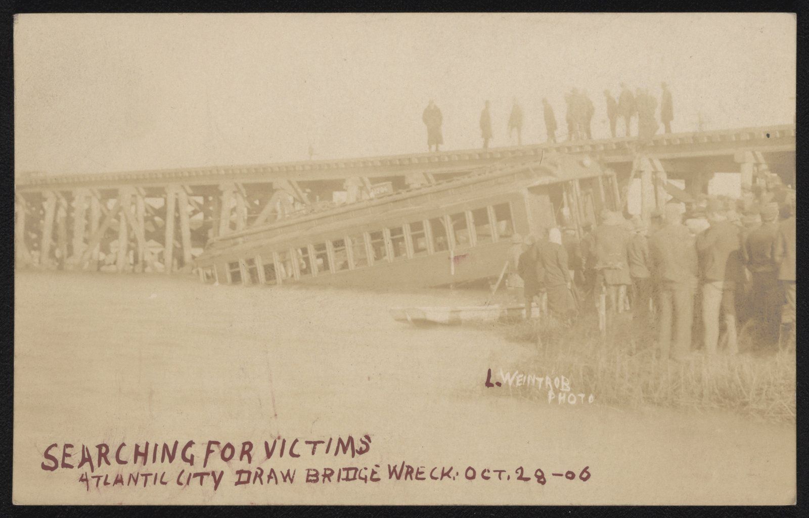 Sepia postcard photograph of a railroad coach lying tilted in shallow water beside a wooden trestle, with men standing on the bridge above and a crowd gathered on the bank, captioned "Searching for victims, Atlantic City draw bridge wreck, Oct. 28-06"