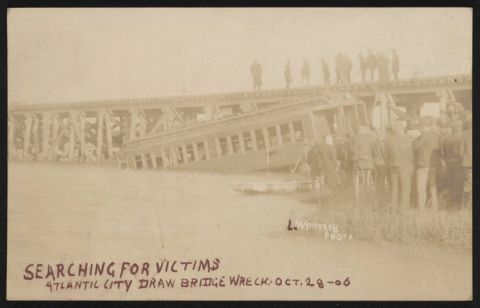 Sepia postcard photograph of a railroad coach lying tilted in shallow water beside a wooden trestle, with men standing on the bridge above and a crowd gathered on the bank, captioned "Searching for victims, Atlantic City draw bridge wreck, Oct. 28-06"