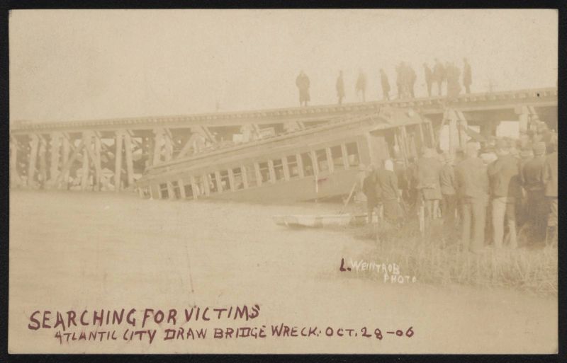 Sepia postcard photograph of a railroad coach lying tilted in shallow water beside a wooden trestle, with men standing on the bridge above and a crowd gathered on the bank, captioned "Searching for victims, Atlantic City draw bridge wreck, Oct. 28-06"