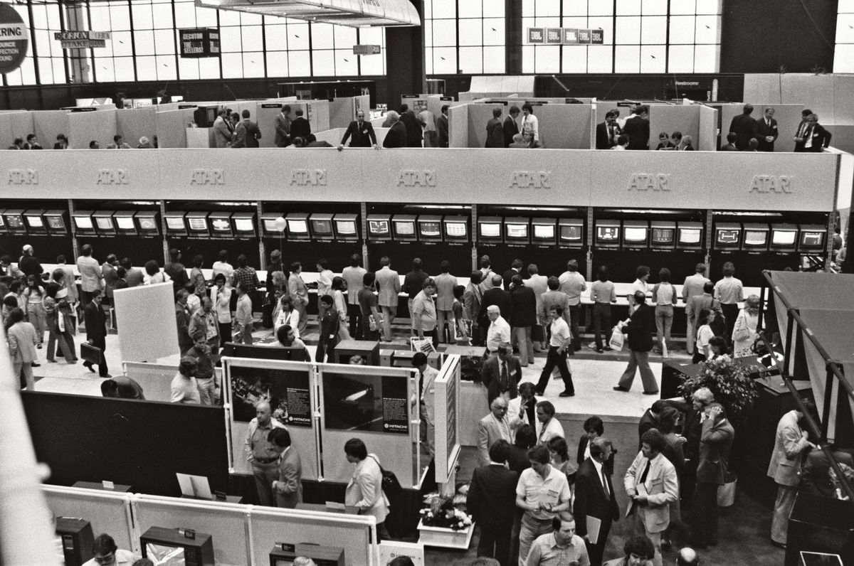 Black and white photo from a balcony over a crowded trade show floor, with a long row of video game screens under repeated Atari signs