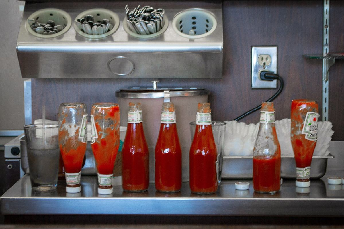 Glass Heinz ketchup bottles lined up on a stainless steel diner counter, some standing upright and full, others turned upside down on top of other bottles to drain