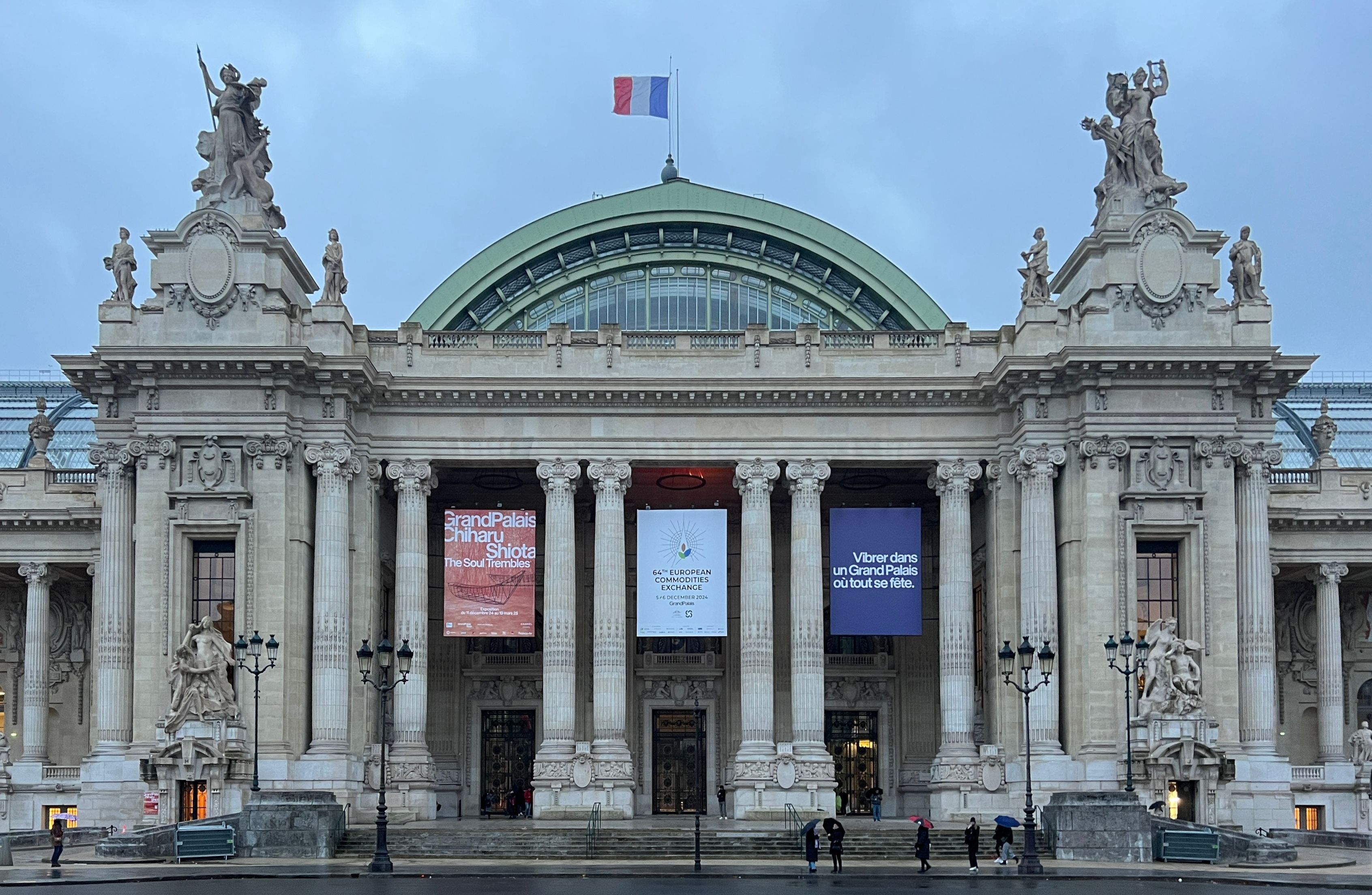 The stone front of the Grand Palais in Paris at dusk, with a green glass dome, columns, statues on the corner pavilions, a French flag on top and three exhibition banners between the columns