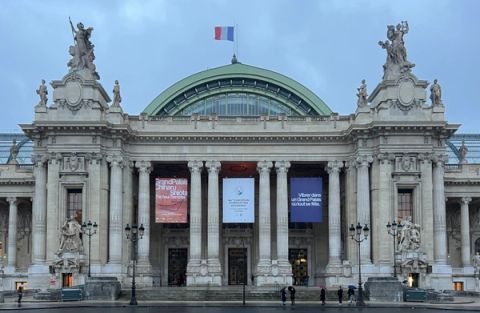 The stone front of the Grand Palais in Paris at dusk, with a green glass dome, columns, statues on the corner pavilions, a French flag on top and three exhibition banners between the columns