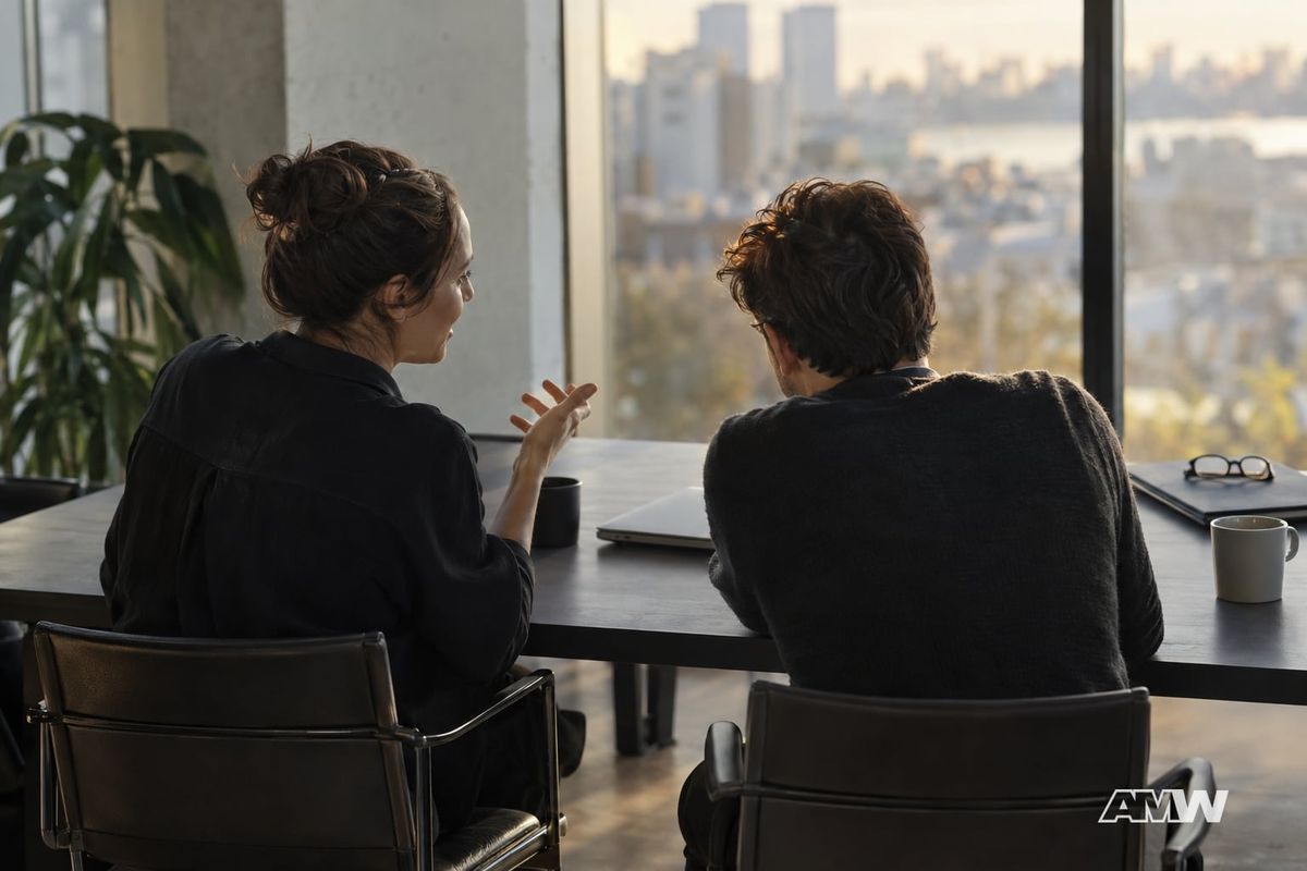 Two colleagues sit at a long office table near a window, gesturing in conversation beside a closed laptop and mugs.