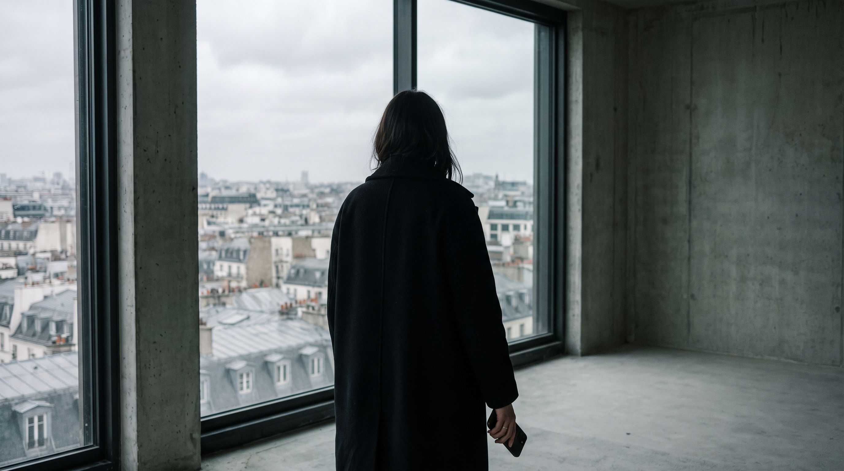 Woman in a black coat looking out over city rooftops from a high window on a grey morning
