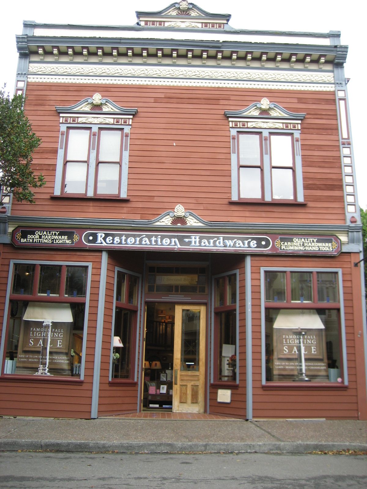 Two-story red wooden Victorian storefront with a Restoration Hardware sign and lighting sale posters in the windows