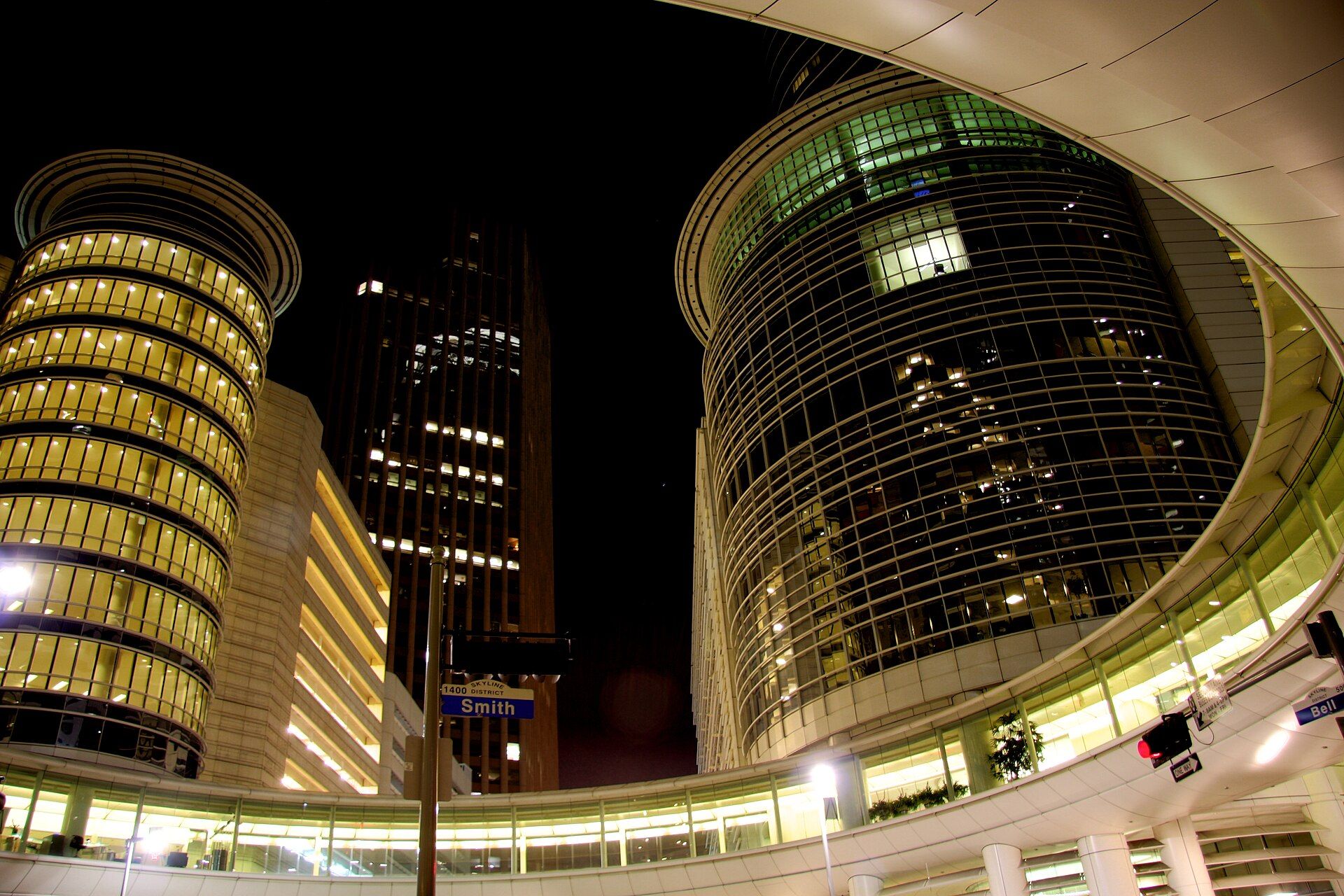 The Enron Complex in downtown Houston at night, a curved glass tower and a round banded tower lit from inside, joined by an elevated curved walkway above a street sign for Smith Street