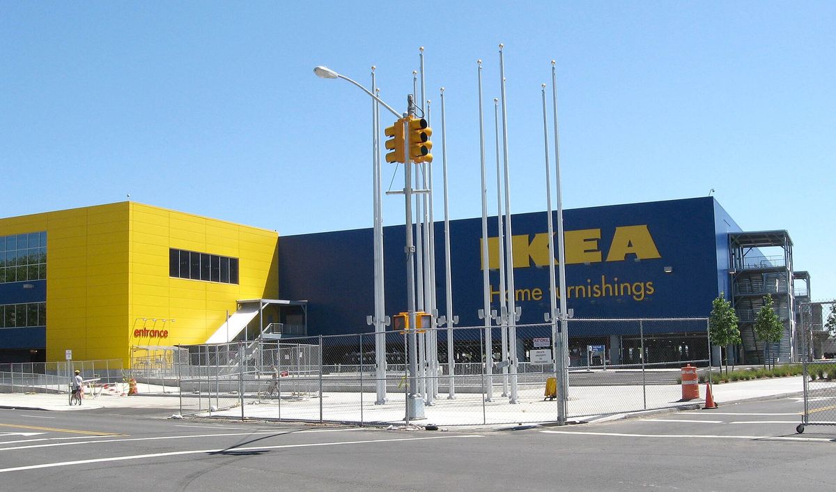 A large blue and yellow IKEA building with the words IKEA Home furnishings on the side, empty flagpoles and a fenced lot in front, under a clear sky