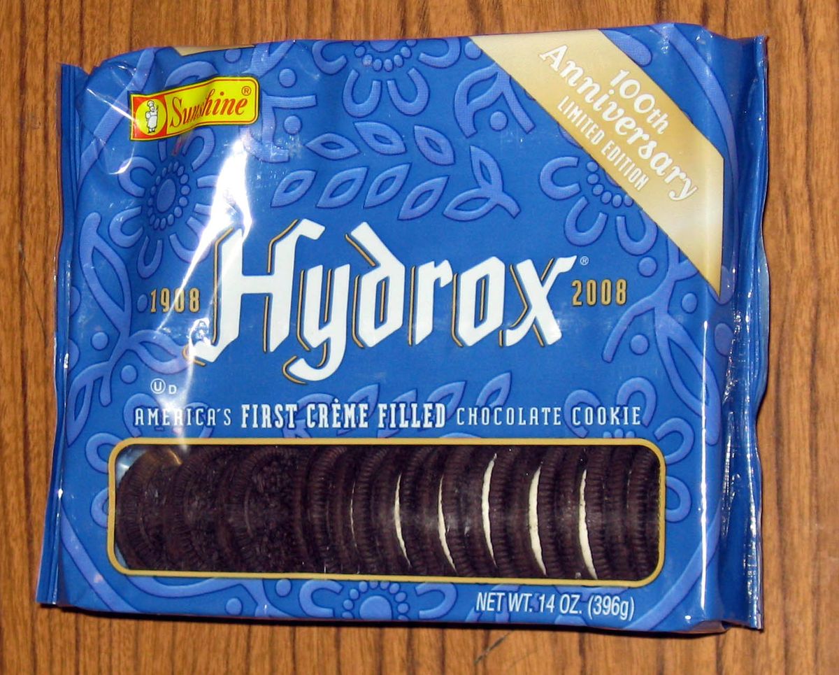 Blue plastic package of Sunshine Hydrox cookies on a wooden table, marked 1908 to 2008, 100th Anniversary Limited Edition, and America's first creme filled chocolate cookie, with a window showing a row of chocolate sandwich cookies