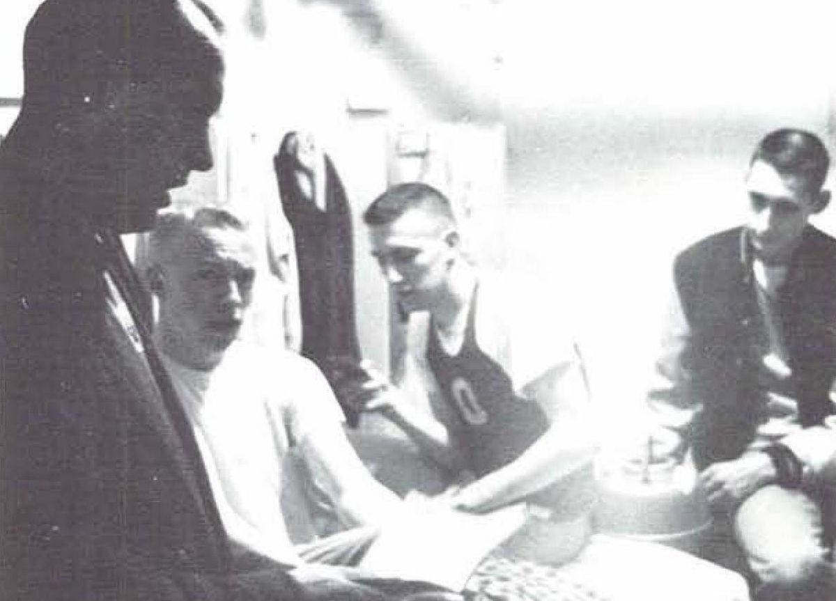 Black-and-white photo of coach Bill Bowerman, in a dark jacket, leaning in to talk with young University of Oregon runners seated in a locker room
