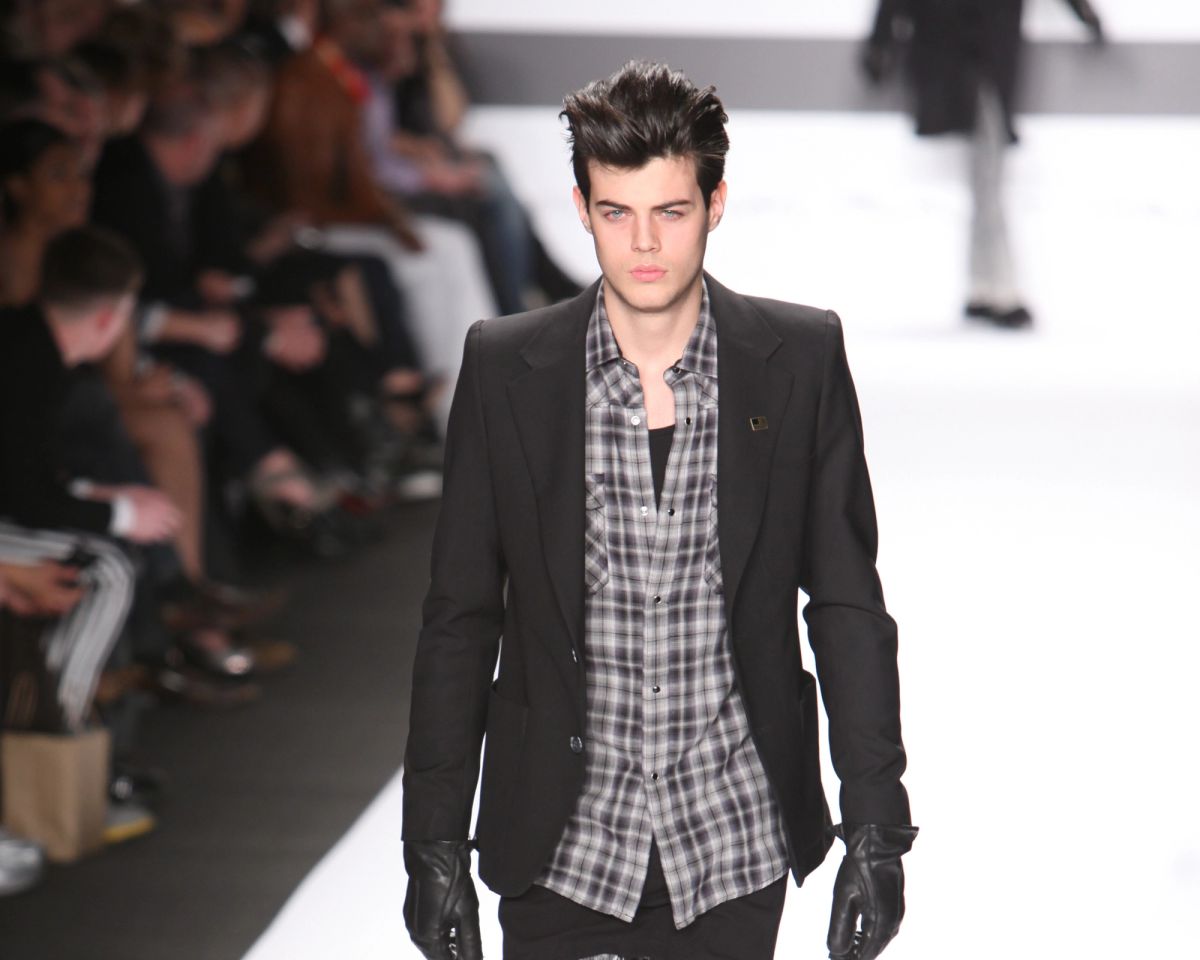A male model in a black blazer, plaid shirt and black leather gloves walks toward the camera on a white runway, with the audience seated along the left side and another model walking away in the background