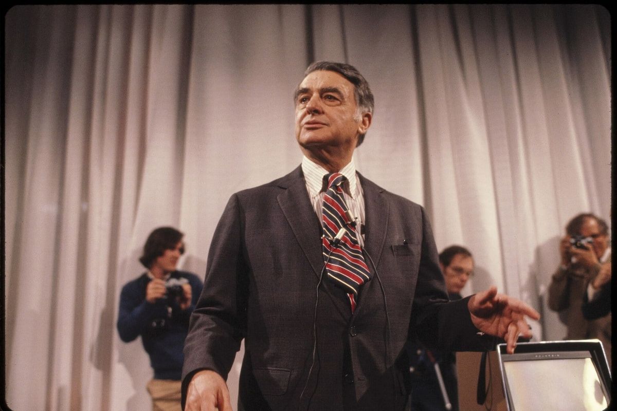 Edwin H. Land in a suit and striped tie speaking at a presentation in front of a curtain background