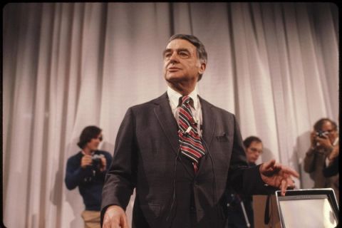 Edwin H. Land in a suit and striped tie speaking at a presentation in front of a curtain background