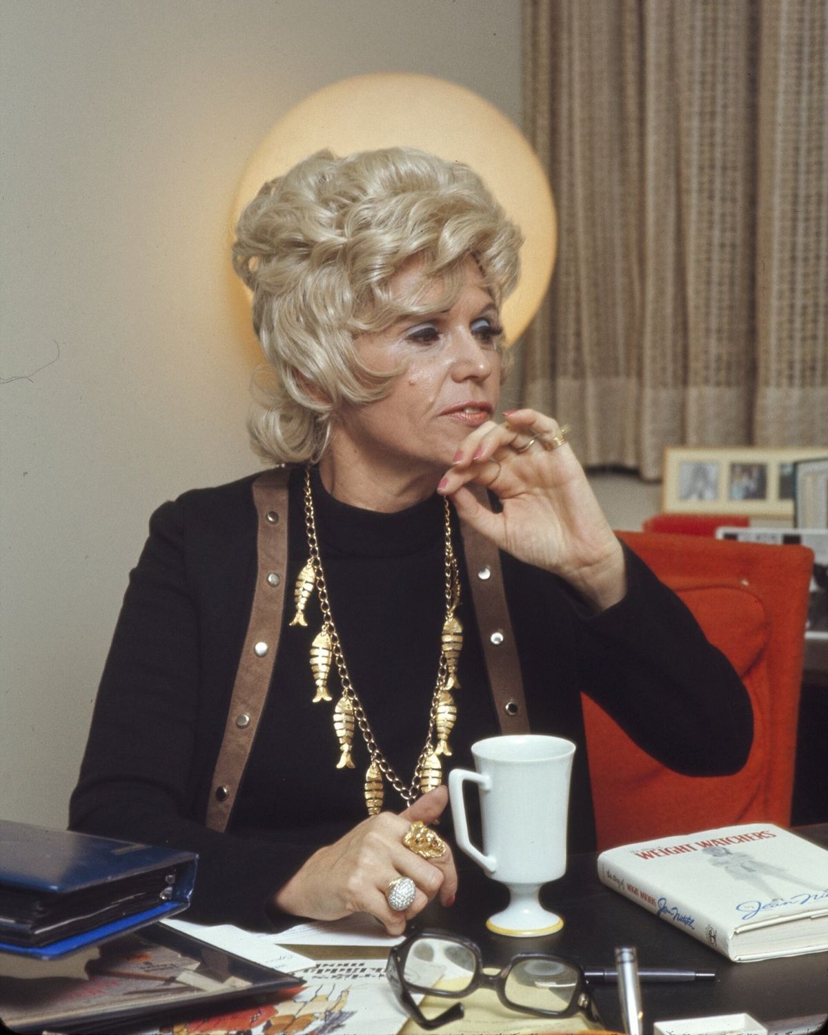 Jean Nidetch, with curled blonde hair and a gold necklace of fish charms, sits at a desk with a white mug and a copy of a Weight Watchers book in front of her