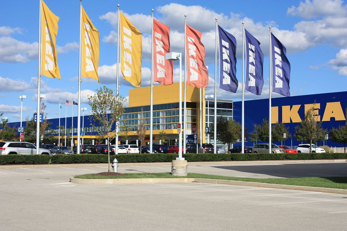 Rows of yellow, orange and blue IKEA banners on flagpoles in front of a blue and yellow IKEA store with a large parking lot under a blue sky