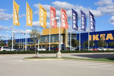 Rows of yellow, orange and blue IKEA banners on flagpoles in front of a blue and yellow IKEA store with a large parking lot under a blue sky