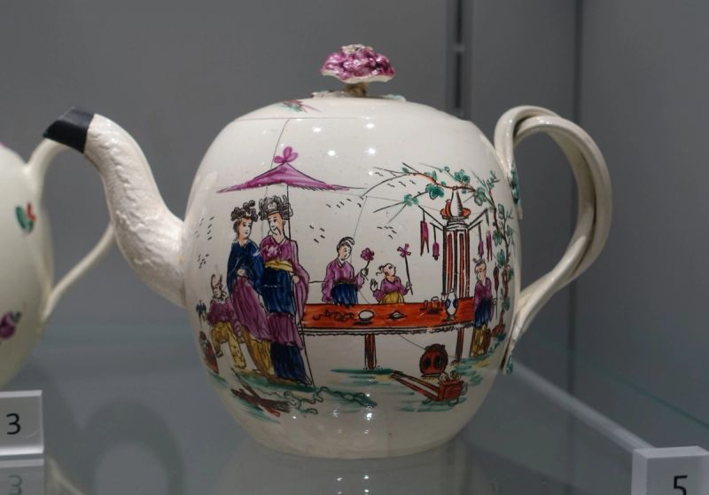 Cream-colored Wedgwood teapot with a twisted handle, painted with figures in long robes under a pink parasol beside a red table, in a museum case